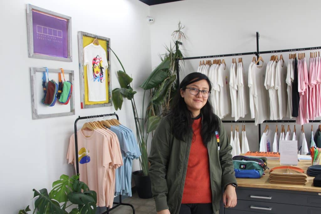 IMG_5151 May Yang stands inside of Flash Flood Print Studios in front of their merch section