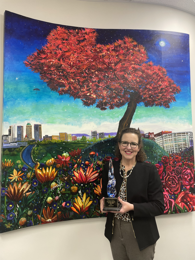 Brittany Littleton holding her BA Chamber award in front of Tulsa to BA painting